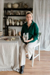 Ceramic artist Kerri of Nioka Clay sits on a barstool at her home studio, holding a ceramic candelabra she crafted.