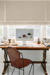 A vintage desk and rattan chair in an office underneath roman blinds.
