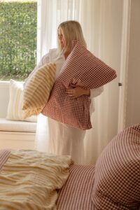 A woman holding two pillows as she styles a bed with Pinot Gingham and Yellow Stripe bed linen.