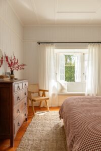 A bed made alongside a Queenslander window seat with white sheer curtains.