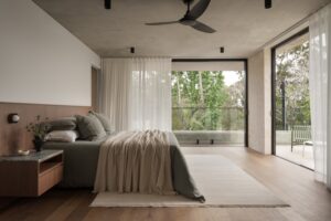 Bedroom with bed covered in olive green linen and a beige throw. The bed sits on a cream rug. Bedroom doors open up to outdoor verandah and bush views.