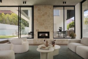 Lounge room looking out to outdoor area, with two armchairs on the left, a lounge on the right, and coffee tables in the centre. Fireplace in the mid-ground and outdoor area in the background.