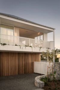 A street view of a modern architectural home, featuring white metal cladding, oak panels, and stone work.