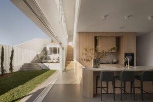 A modern architectural kitchen with sliding doors that open to an outdoor area, featuring brass finishings, upholstered bench stools, and oak tones.