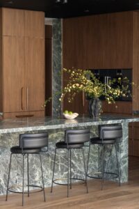 A kitchen with marble bench top and walnut cabinetry. A vase with greenery and yellow flowers sits atop the bench, to the right of a small ceramic fruit bowl filled with limes. Three black leather stools positioned at the bench.