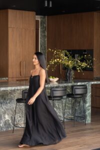 A kitchen with marble bench top and walnut cabinetry. A vase with greenery and yellow flowers sits atop the bench, to the right of a small ceramic fruit bowl filled with limes. Three black leather stools positioned at the bench. A woman in a black dress is walking across the shot toward the left.