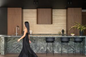 A kitchen with marble bench top, gunmetal tapware, and walnut cabinetry. A woman in a black dress is walking across the shot toward the left.