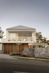 A street view of a modern architectural home, featuring white metal cladding, oak panels, and stonework.