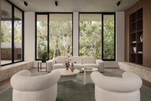 Sunken lounge room with two white armchairs in the foreground, coffee tables in the centre, and a three-seater sofa against the large windows with bush views. Walnut cabinetry on the right.