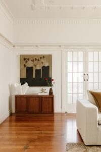 High ceilings with a painters rail, crown moulding, and plastered ceiling detail are pictured in the living area of a renovated Queenslander. A painting by artist Giorgia Bel hangs on the wall above a mid-century cabinet topped with fresh flowers, vintage ceramics, an a blown glass lamp.
