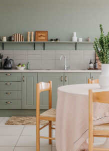 Looking past a round table surrounded by rush chairs to a kitchen painted sage green. There are black Smeg appliances on the bench as well as handmade ceramics and eclectic pieces along an exposed shelf.