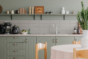Looking past a round table surrounded by rush chairs to a kitchen painted sage green. There are black Smeg appliances on the bench as well as handmade ceramics and eclectic pieces along an exposed shelf.