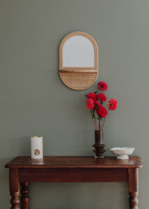 A rattan mirror hanging on a sage green wall over an antique timber console table with turned legs. Red dahlias sit on it alongside handmade ceramics while rattan baskets sit underneath.