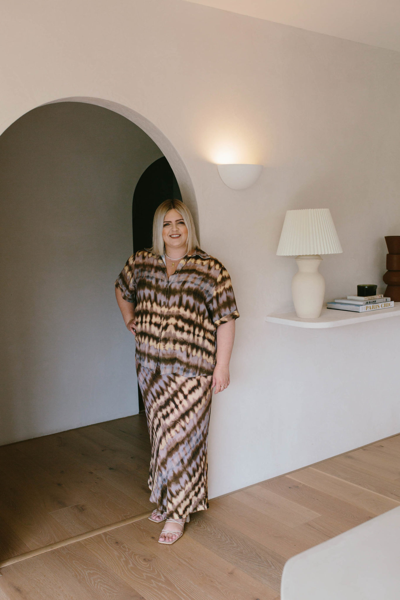 Hayley Jenkin stands in an archway with a lamp and objects on a shelf to her right.